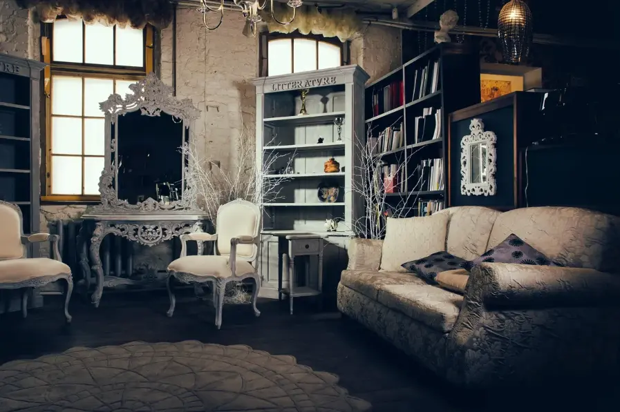Vintage furniture showroom with ornate mirror, upholstered chairs, shelving displaying decorative objects, and worn industrial brick walls lit by large windows