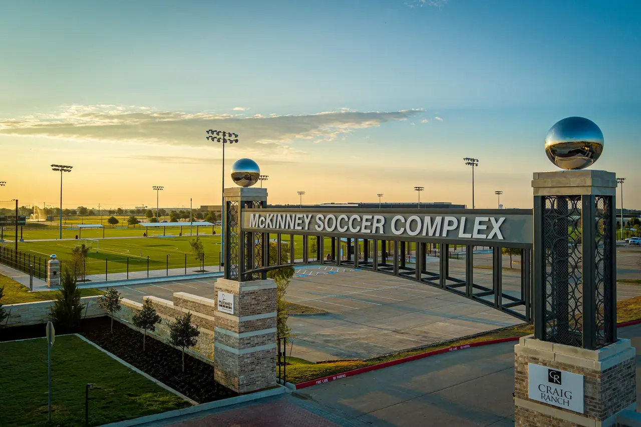 Entrance to soccer complex with illuminated fields at dusk, modern signage, and manicured grass grounds under evening sky