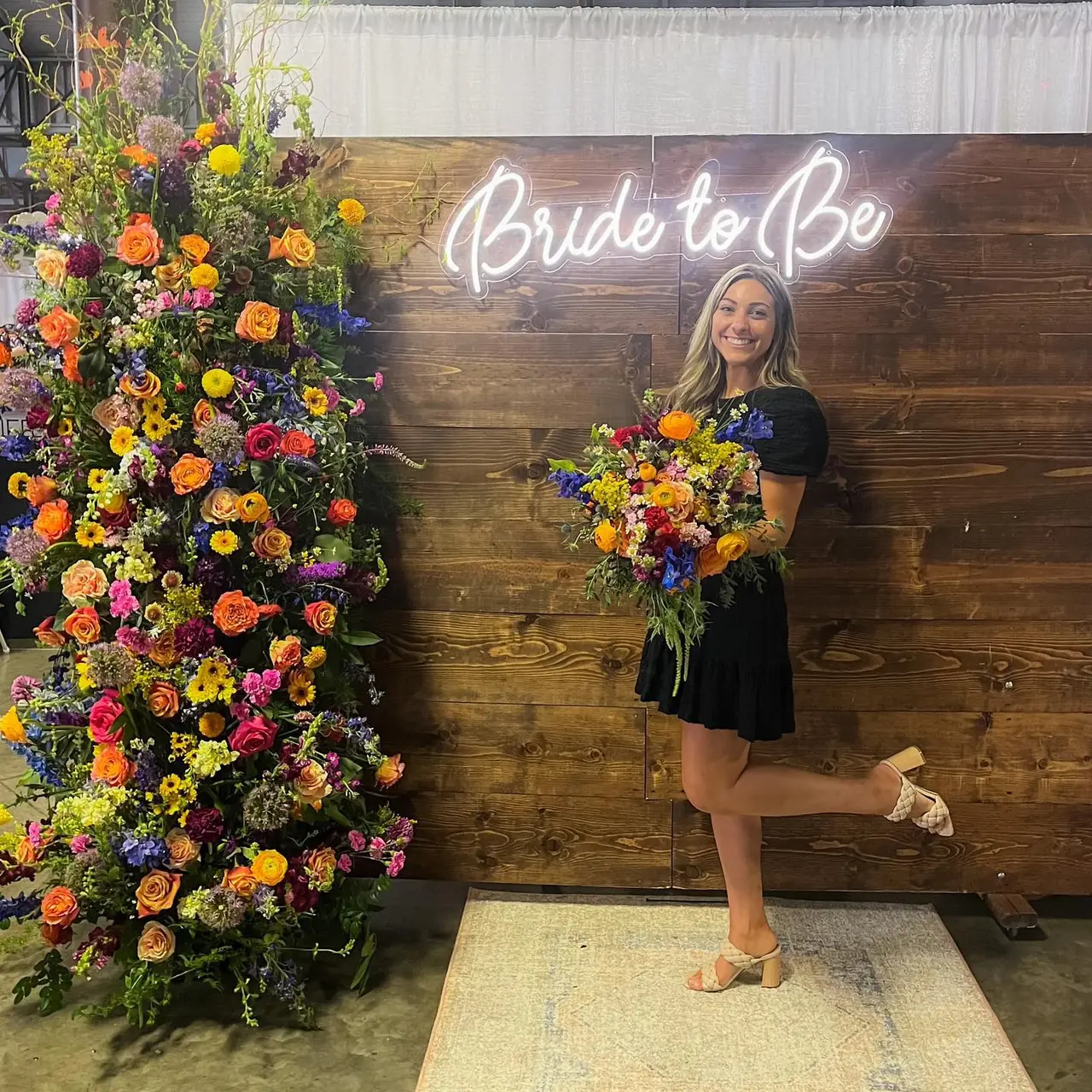 Woman holding colorful flower bouquet standing in front of neon "Bride to Be" sign with vibrant floral wall display