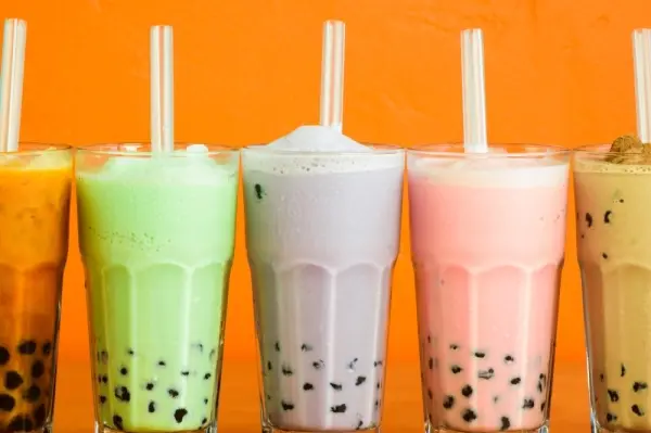 Five colorful bubble teas with tapioca pearls and straws against an orange background, including amber, green, white, pink, and yellow drinks