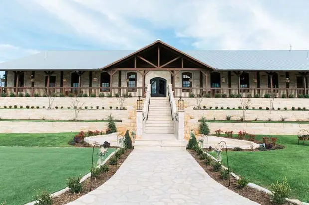 Large rustic barn with stone and wood exterior, wide front porch, central entrance with stairs, manicured lawn and landscaping
