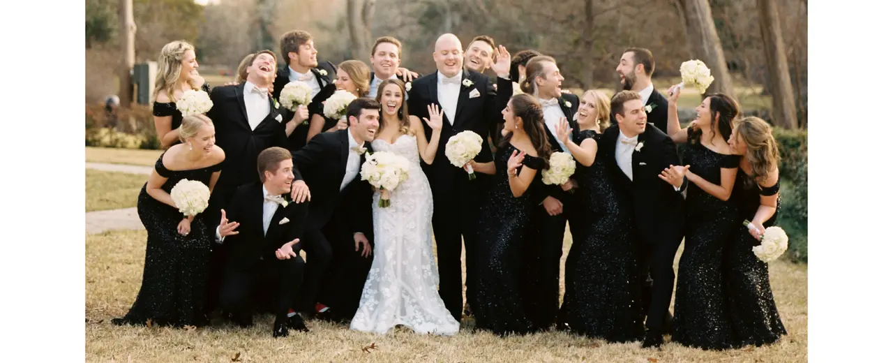 Bride in white wedding dress surrounded by wedding party in black formal attire holding white bouquets outdoors