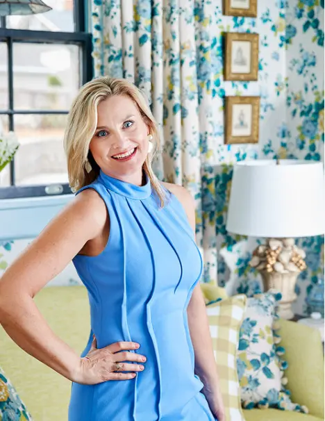 Woman in blue dress posing in living room with blue and teal floral curtains and gold-framed artwork