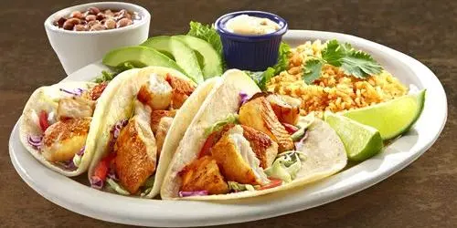Two crispy tacos with shredded chicken, sliced avocado, rice, beans, and lime wedges on a white plate