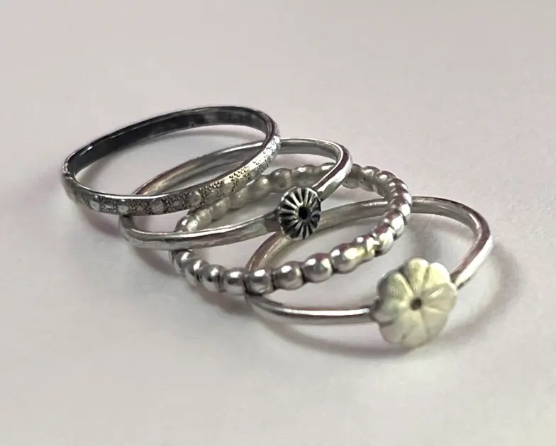 Four silver rings with decorative details including beaded textures and small floral charms, arranged in a stacked composition
