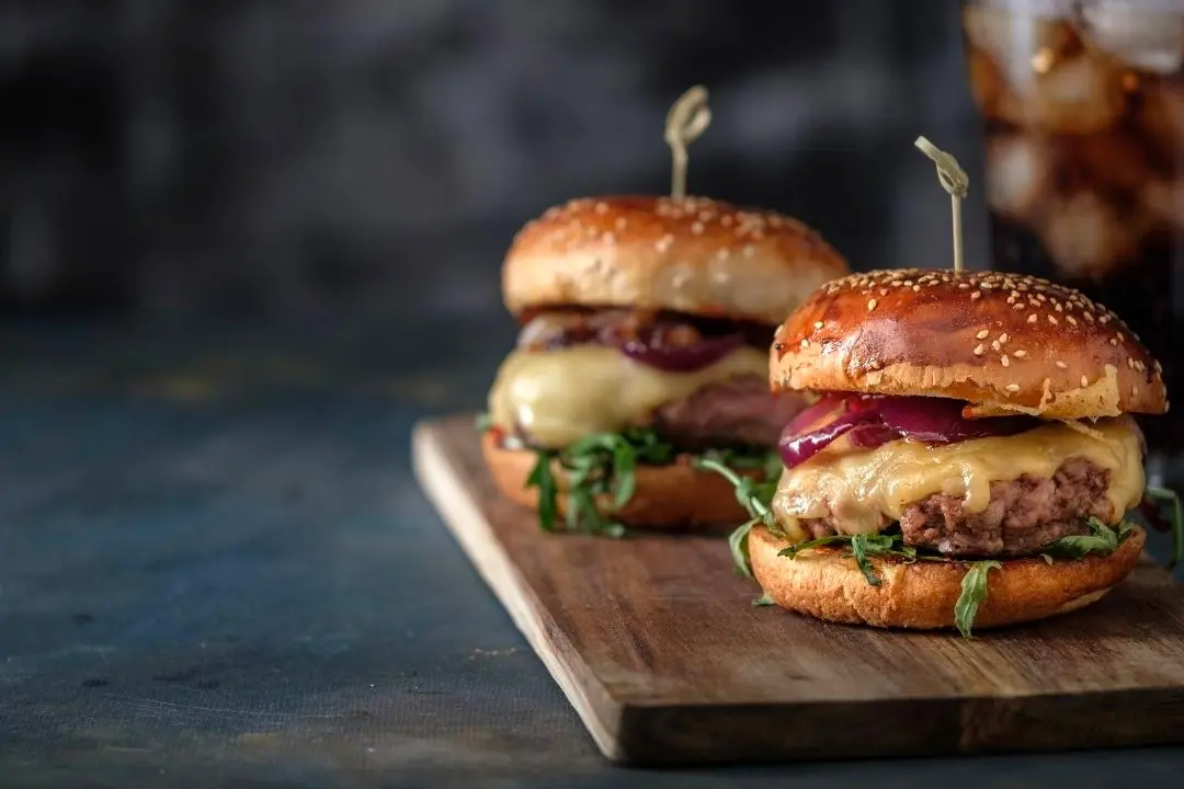 Two gourmet burgers with sesame seed buns, melted cheese, red onion, and fresh greens on a wooden board with toothpick picks