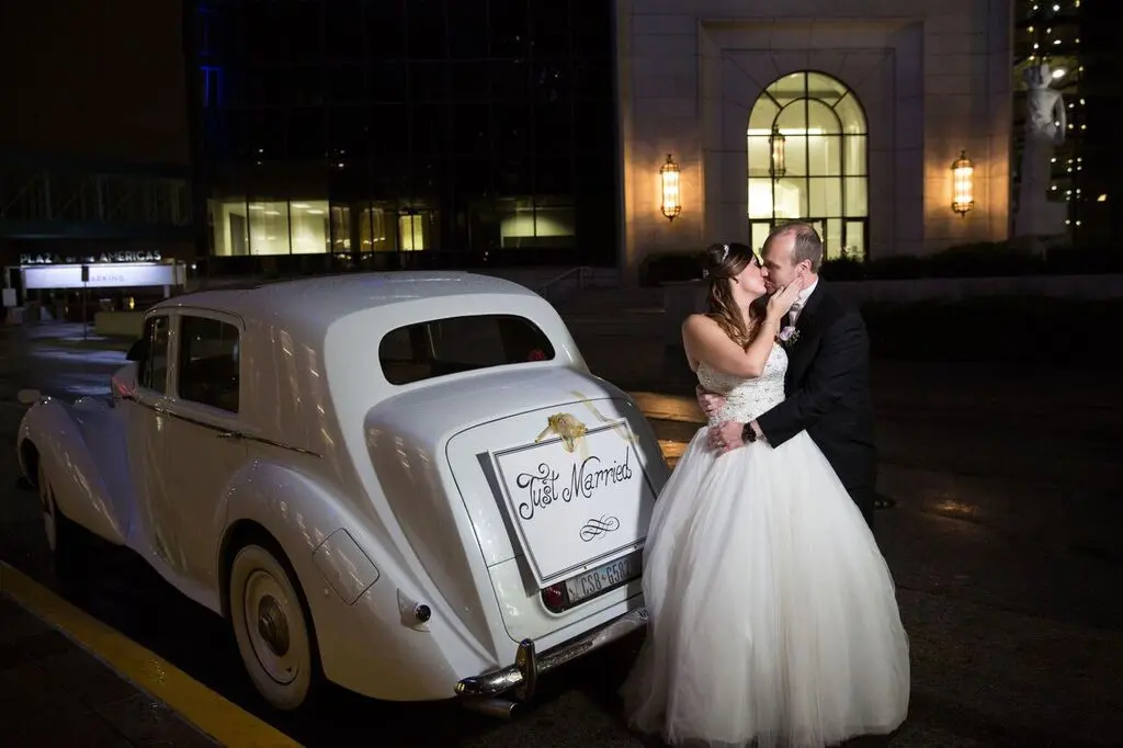 Bride and groom posing together beside a vintage white wedding car at night with illuminated building in background