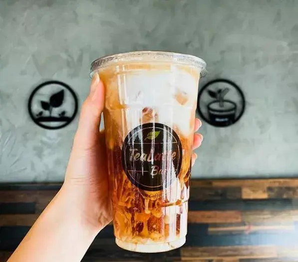 Hand holding a clear cup of iced tea with boba pearls and branded logo, against a gray wall with decorative icons
