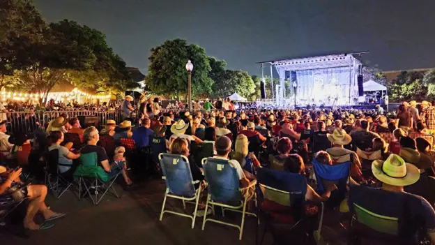 People sitting on lawn chairs in a parking lot listening to music on a stage