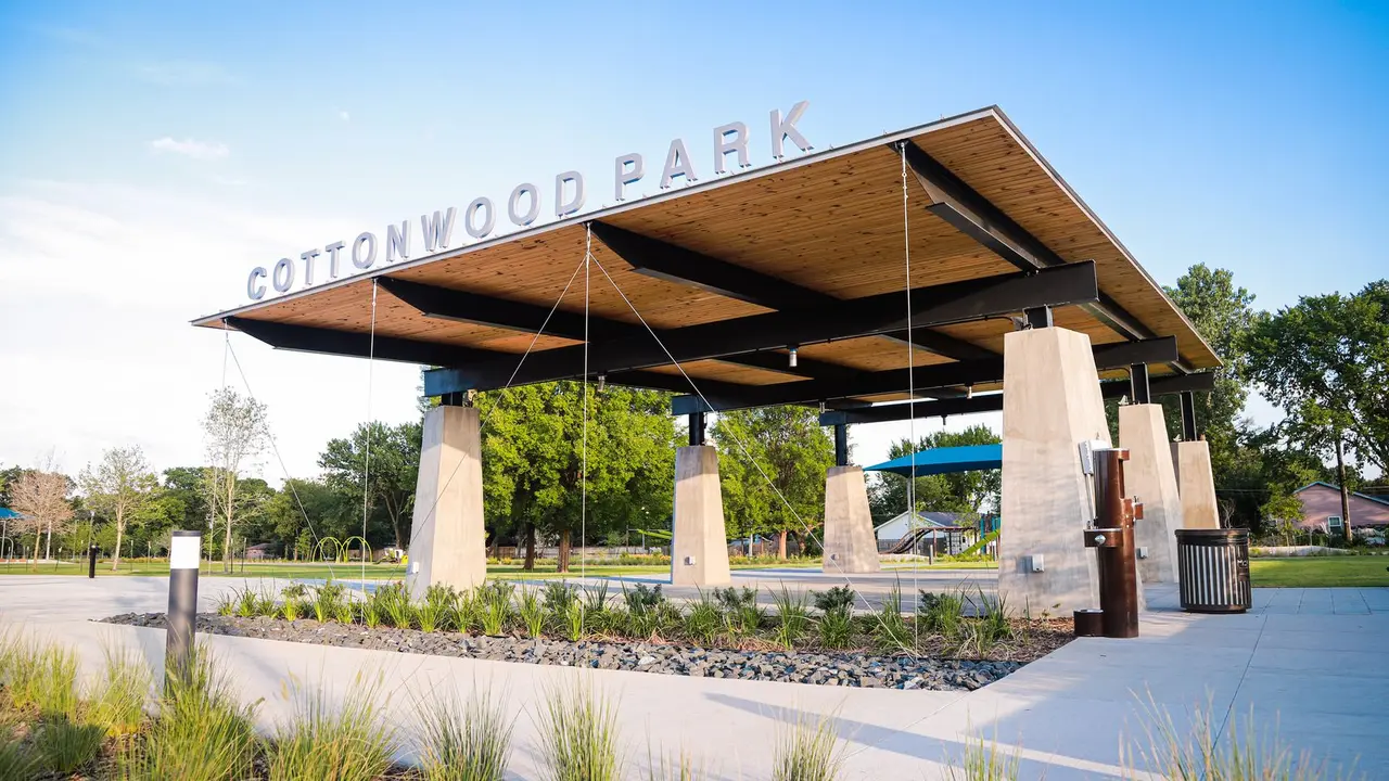 Modern pavilion with wooden roof and concrete pillars at neighborhood park, surrounded by landscaping and trees under clear sky