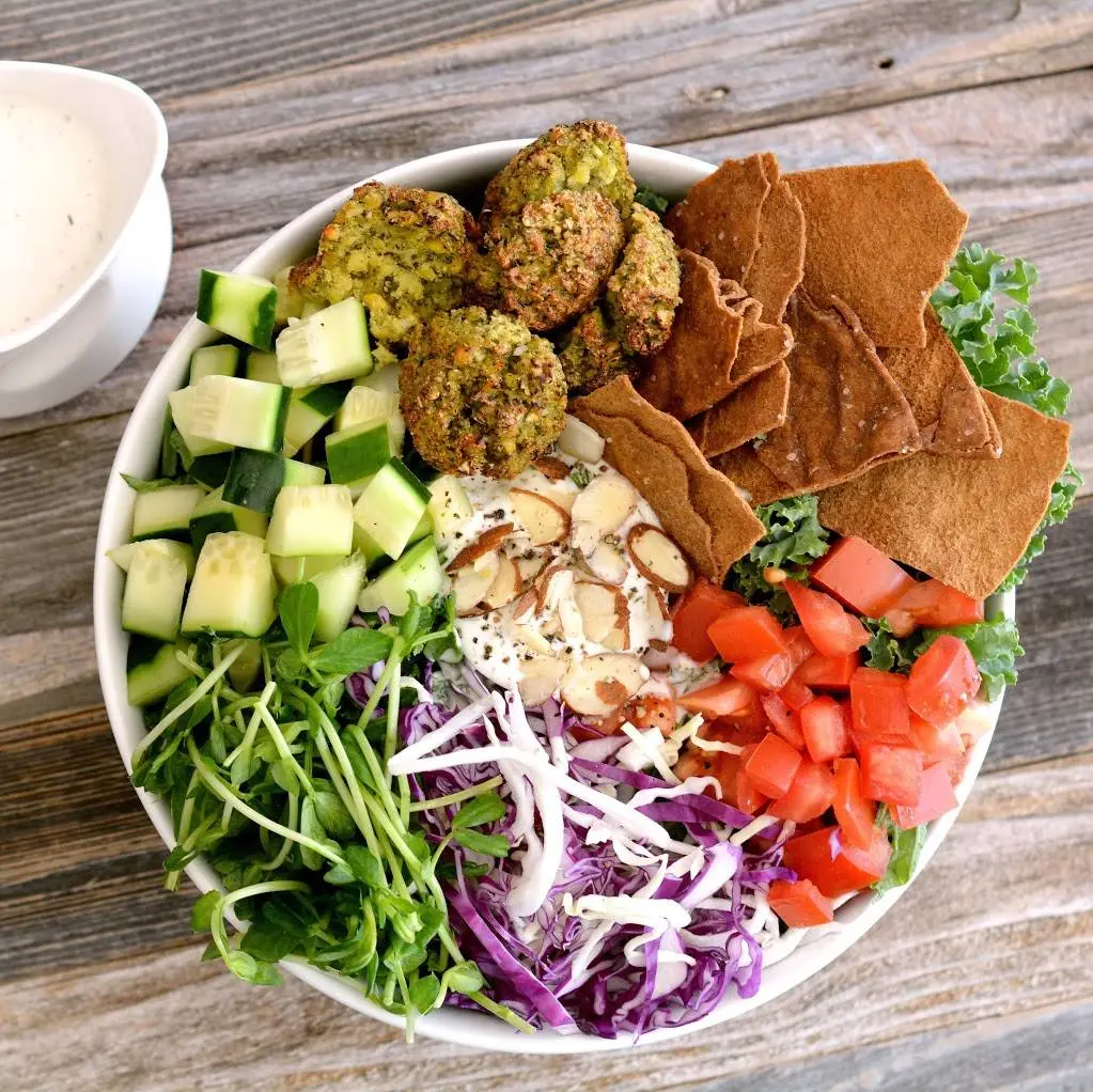 White bowl filled with chopped vegetables, falafel, pita chips, greens, and a white dipping sauce on wooden surface