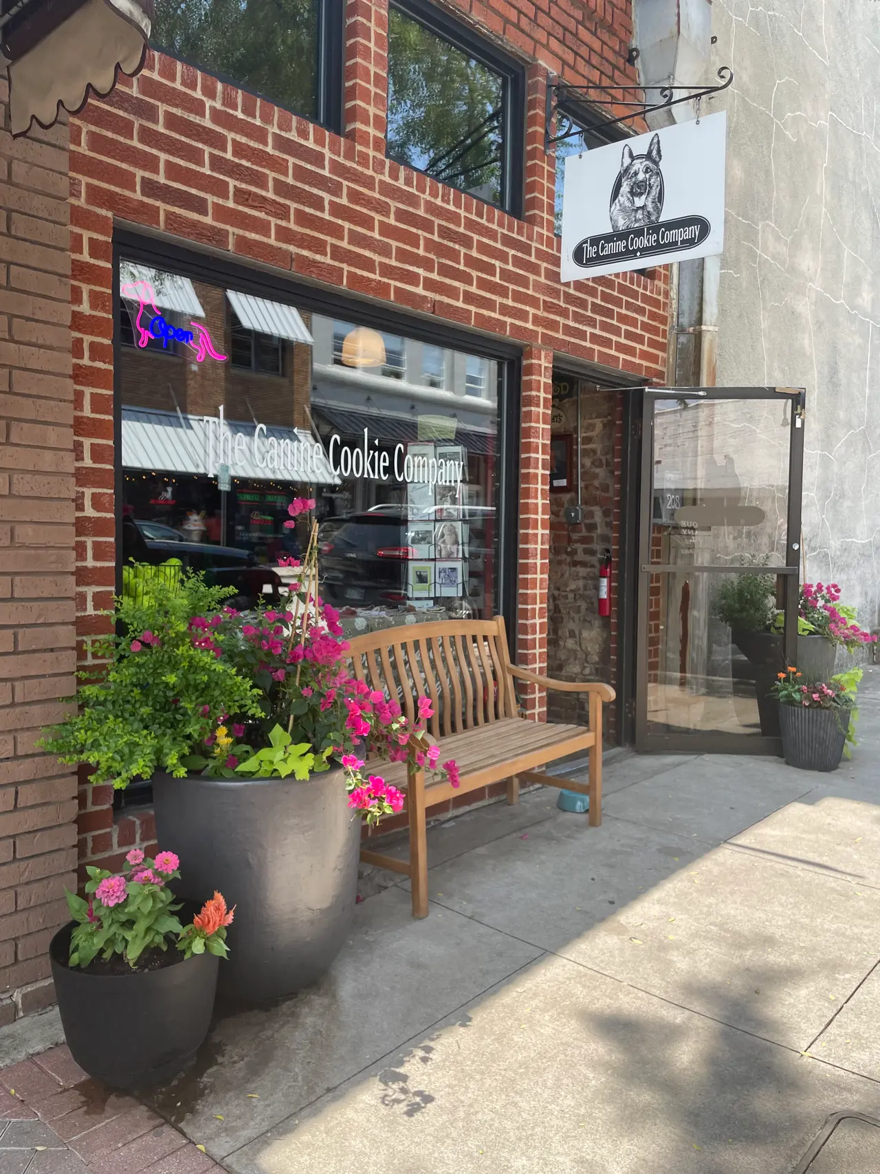 Brick storefront with wooden bench, large planters of pink flowers, and glass door entrance on sidewalk