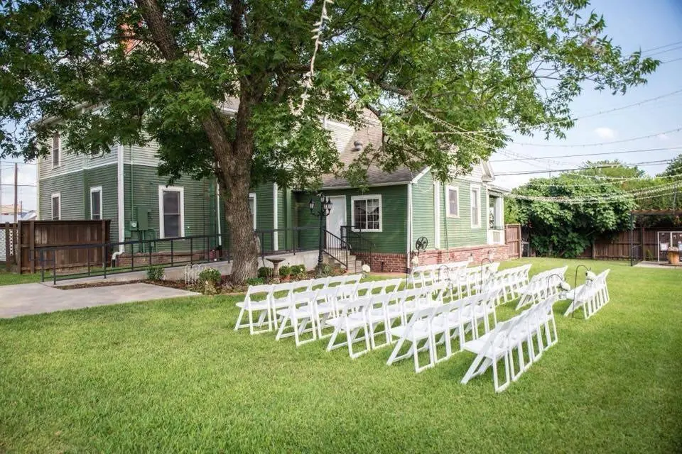 White folding chairs arranged on a lawn facing a green historic house with large shade tree