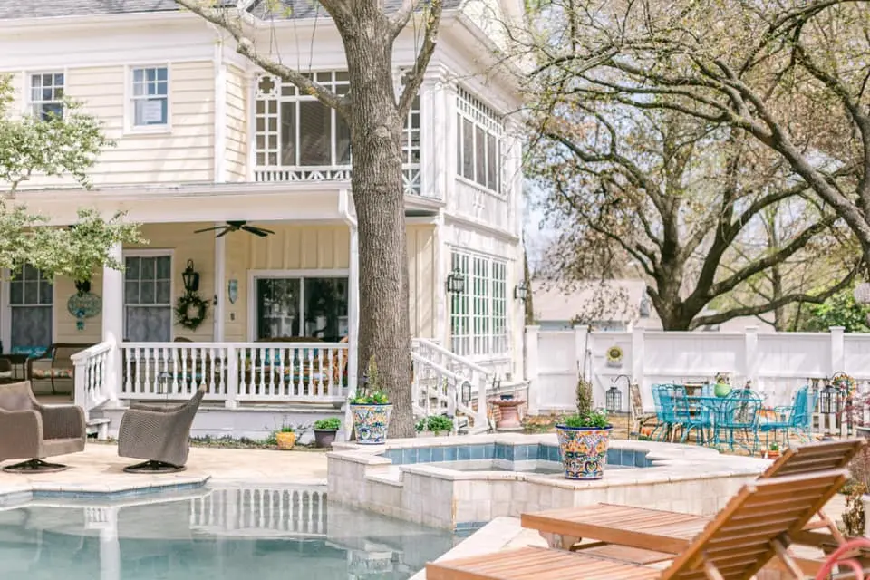 Two-story cream-colored house with wraparound porch overlooking swimming pool and blue lounge chairs, large tree in foreground