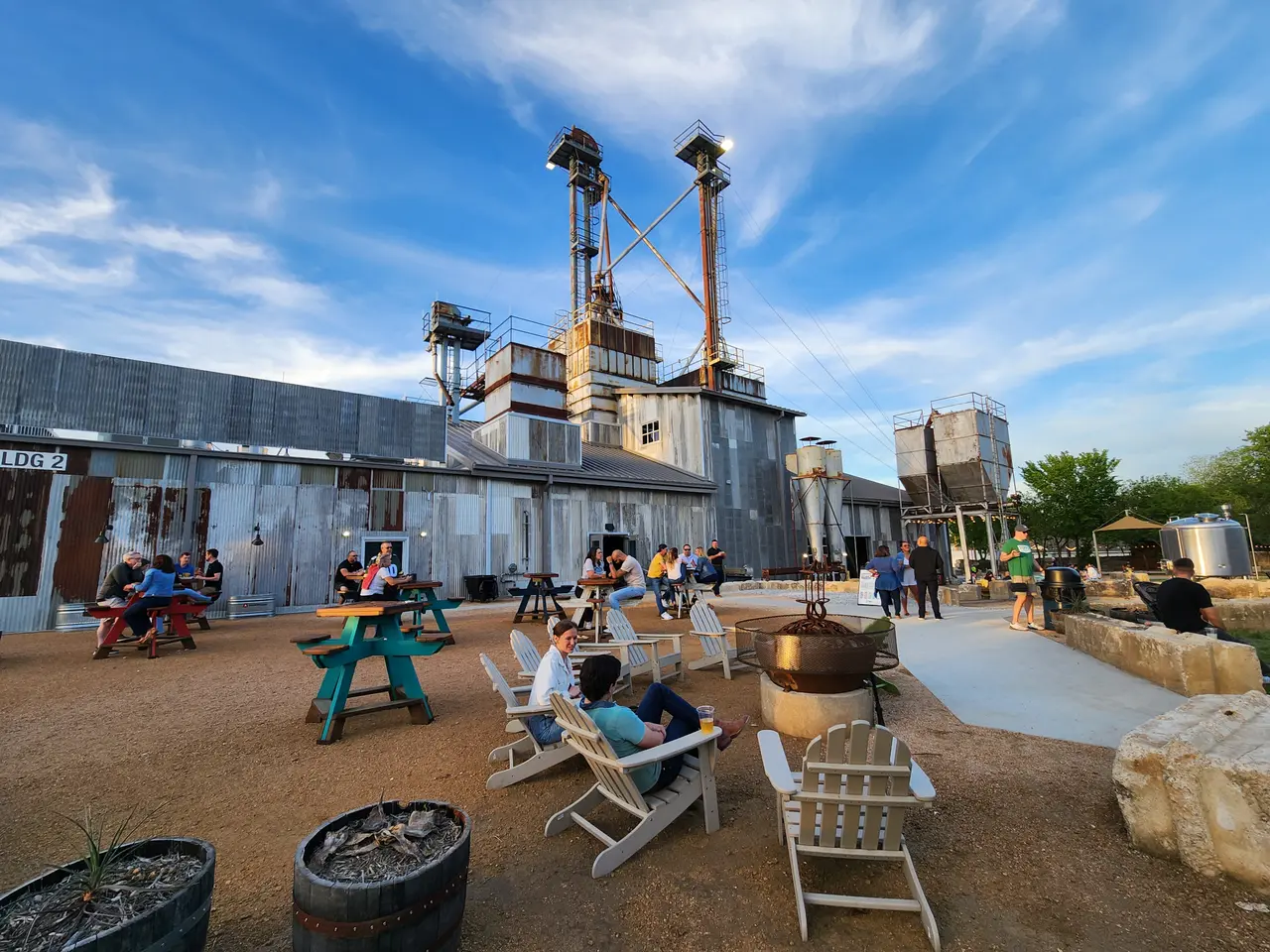 Industrial brewery buildings with tall metal towers, outdoor seating area with picnic tables and lounge chairs on dirt ground, visitors scattered throughout