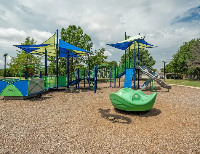 Modern playground with blue shade structures, climbing equipment, and green play features on mulch ground surrounded by trees