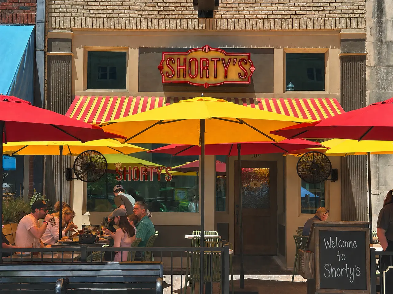 Storefront with red and yellow striped awnings and umbrellas, customers seated at outdoor tables, brick building facade