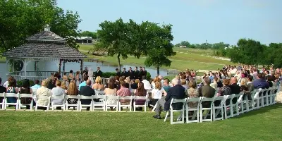 Outdoor wedding ceremony with guests seated in white chairs on a lawn, gazebo structure and tree-lined landscape in background