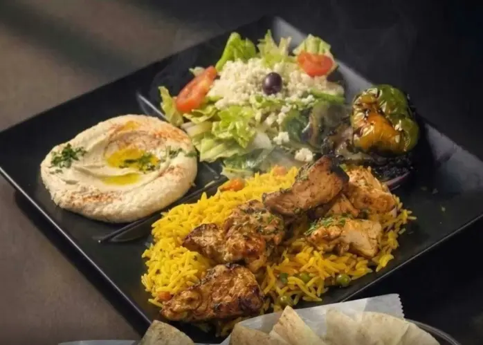 Plate with hummus, grilled chicken kebab on yellow rice, fresh salad, and roasted vegetables on black plate