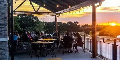 Diners seated at tables on a covered patio at sunset with trees and water visible in the distance