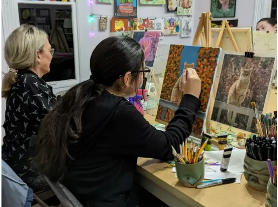 Two people working on artwork at a studio table with easels, art supplies, and drawings displayed on the wall behind them