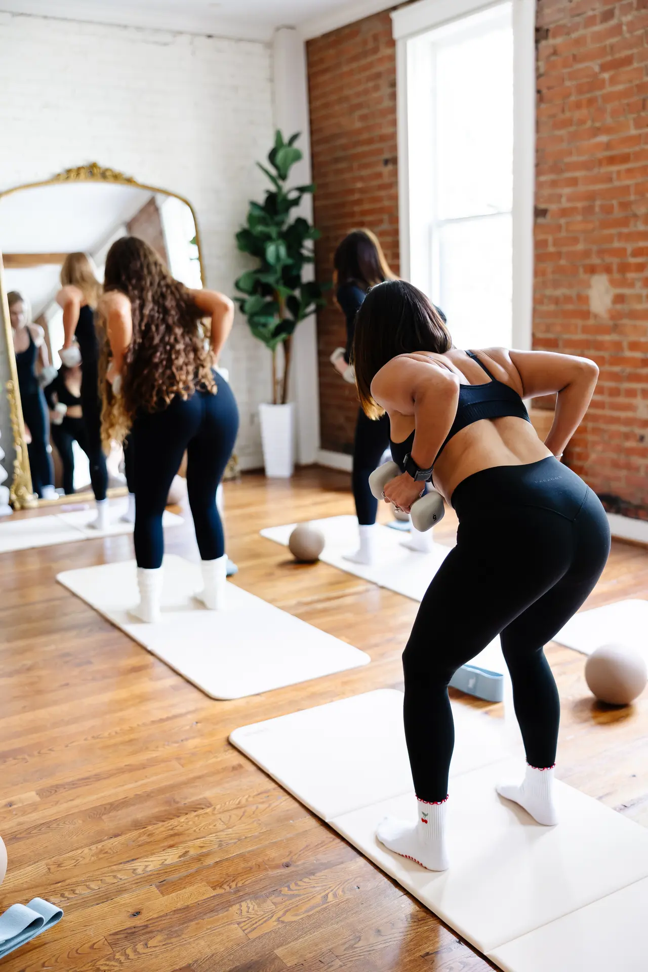 Women doing Pilates exercises on mats in a brick-walled studio with mirrors and potted plants