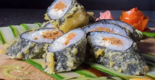 Sushi rolls topped with sesame seeds and filled with avocado and orange roe, arranged on green strips with pickled ginger and red pepper garnish