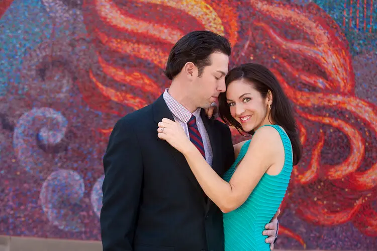 Couple posing together in front of ornate red and blue patterned backdrop, man in dark suit and woman in turquoise sleeveless top smiling at camera