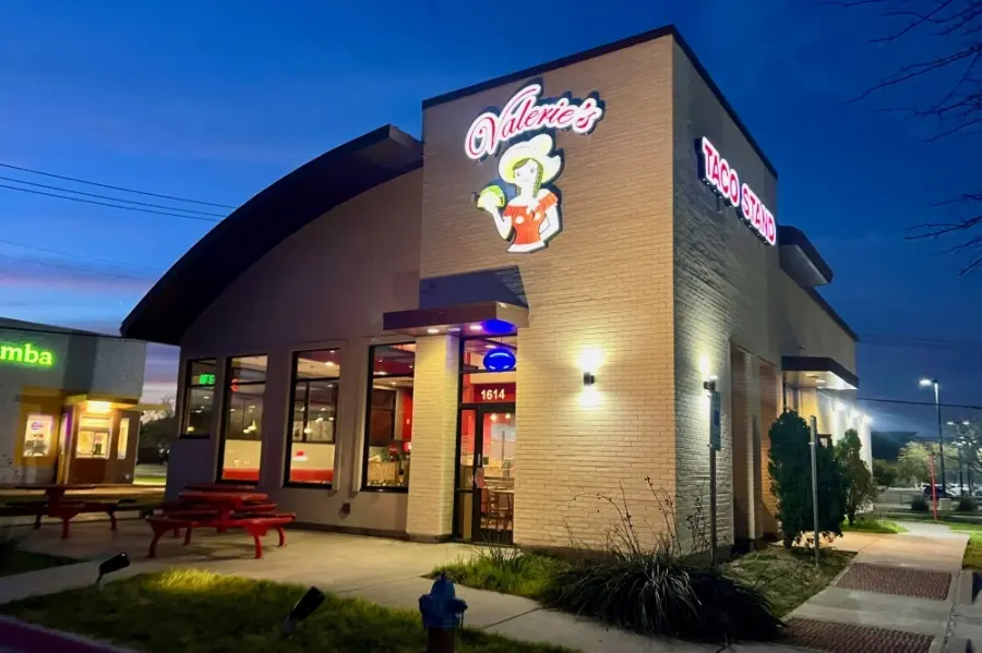 Brick restaurant building with illuminated signage at dusk, storefront windows, and red bench seating outside
