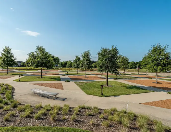 Spacious park with mature trees, curved walking paths, benches, and mulched planting beds under clear blue sky