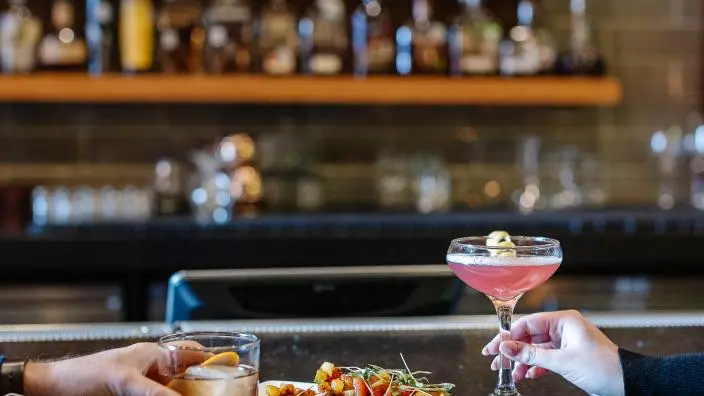 Two hands holding cocktails at a bar counter with appetizers, shelves of liquor bottles blurred in background