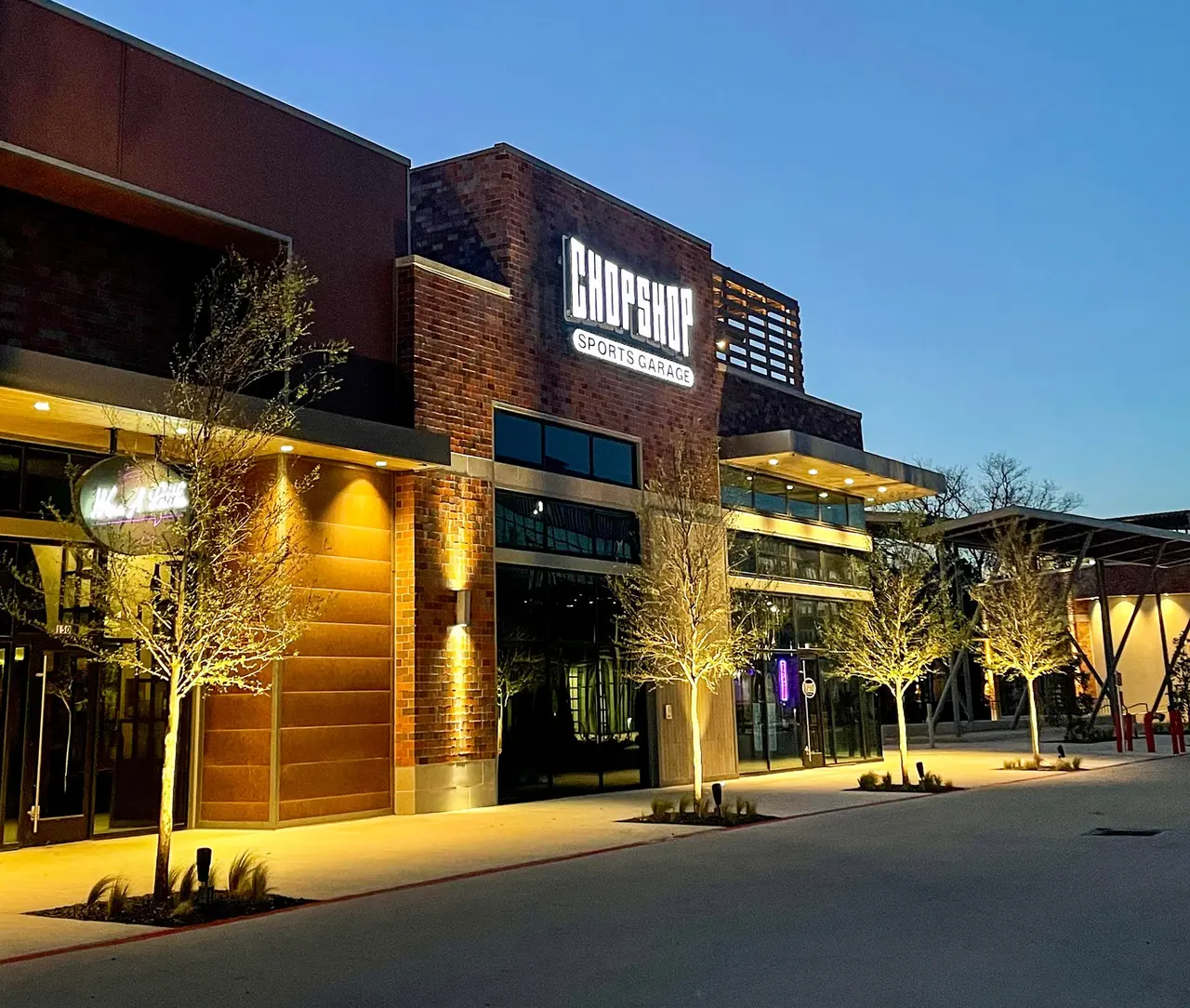 Modern restaurant exterior with brick and wood facade, illuminated by warm yellow lighting at dusk, with young trees and clear blue sky above