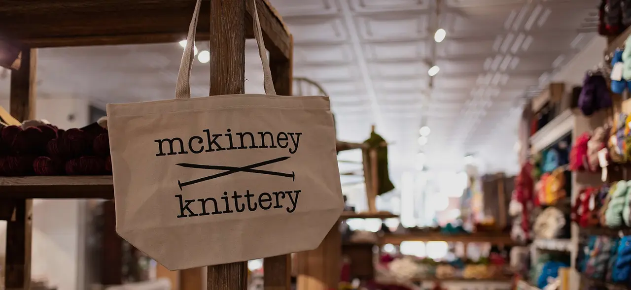 Canvas tote bag with "mckinney knitttery" text hanging in yarn shop interior with colorful merchandise visible in background