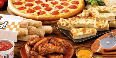 Pepperoni pizza, breadsticks, and fried chicken arranged on a wooden surface with dipping sauces
