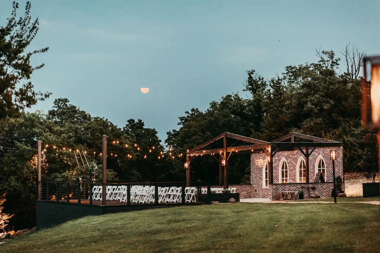 Twilight view of outdoor event space with illuminated wooden pergolas, string lights, and brick building surrounded by trees and lawn