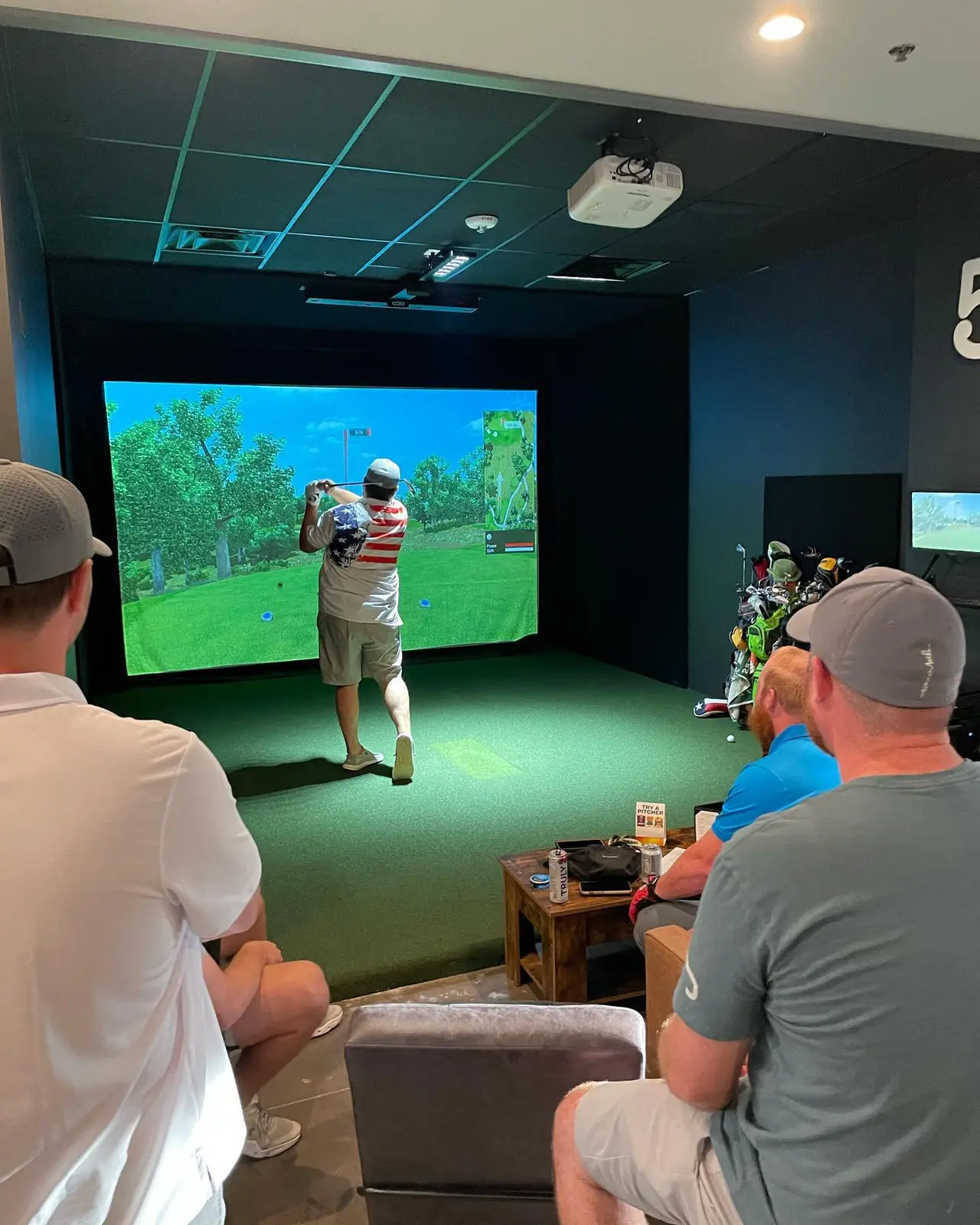 Golfer swinging at indoor simulator with large screen displaying fairway while spectators watch from seating area