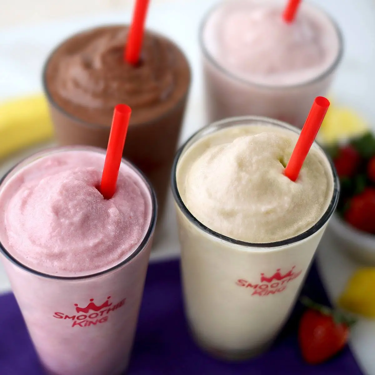 Four blended smoothie drinks with red straws in clear cups, including pink, chocolate, and vanilla varieties
