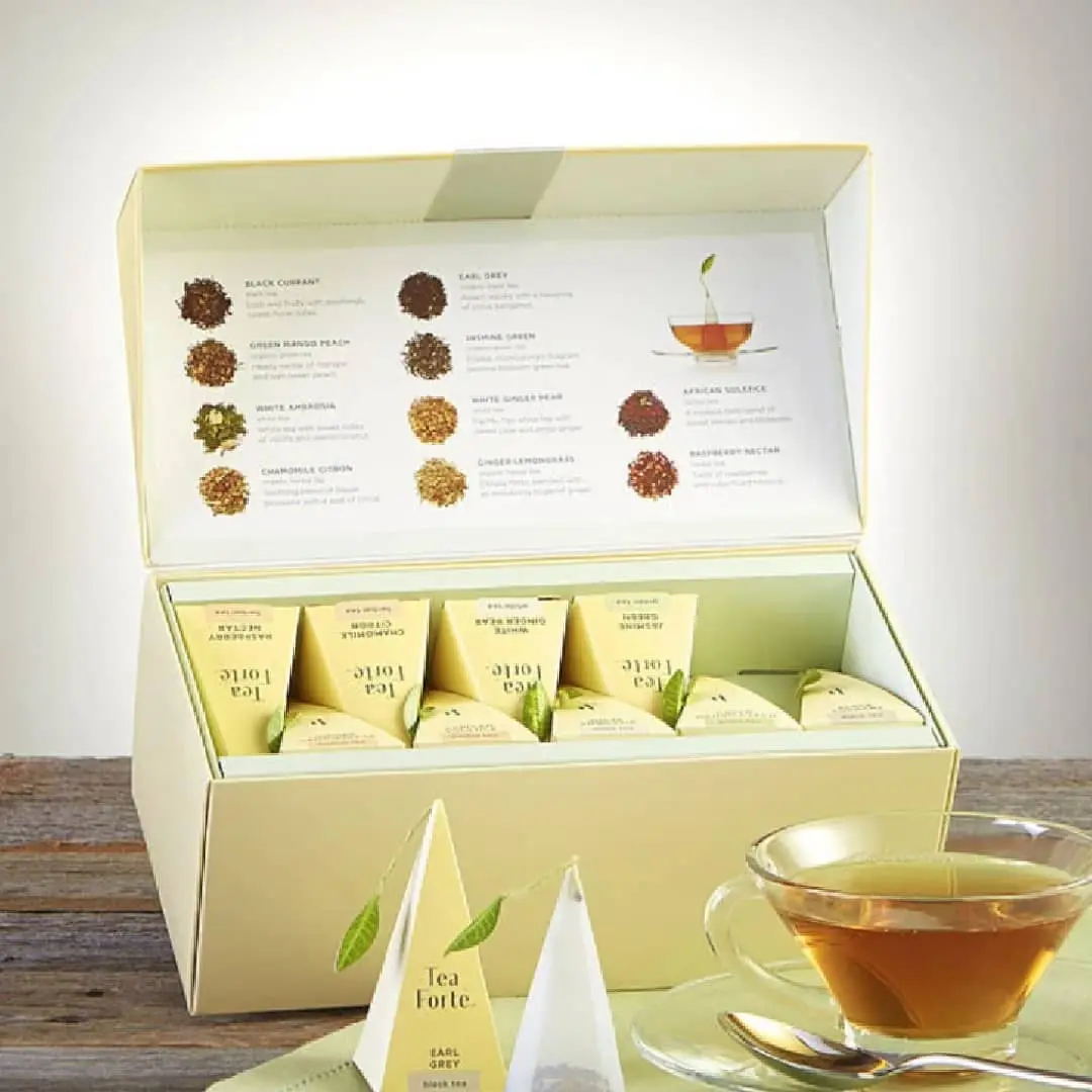 Open gift box with tea sachets, flavor guide showing dried herbs and tea leaves, and brewed tea in glass cup with saucer
