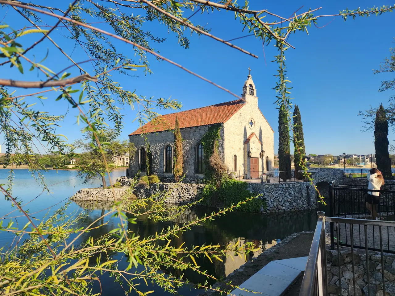 Stone chapel with terracotta roof beside calm water, cypress trees framing the scene, clear blue sky above