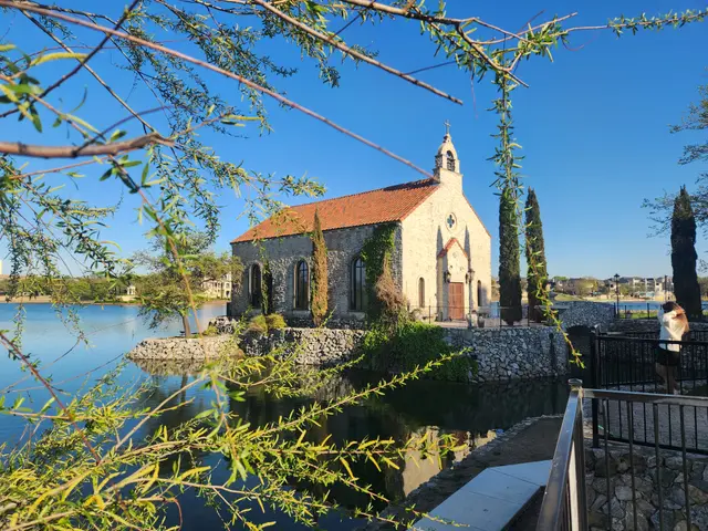 Stone chapel with terracotta roof beside calm water, cypress trees framing the scene, clear blue sky above