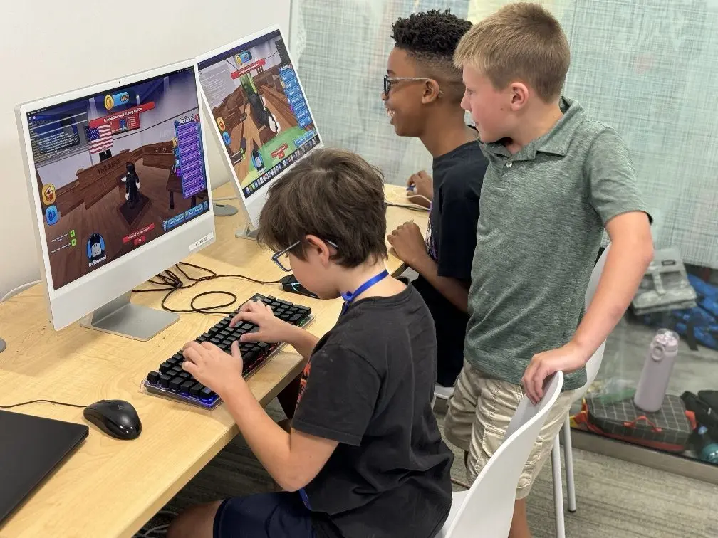 Three young people gathered around a desk, one seated at a computer with a game displayed on screen, two standing beside watching