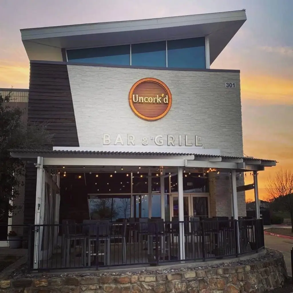Modern brick building with large circular logo and bar & grill signage, illuminated at sunset with outdoor seating area