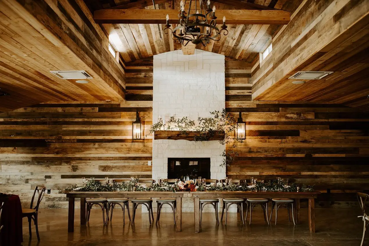 Rustic wooden barn interior with long dining table, fireplace with white mantel, exposed beam ceiling, and floral garland decorations