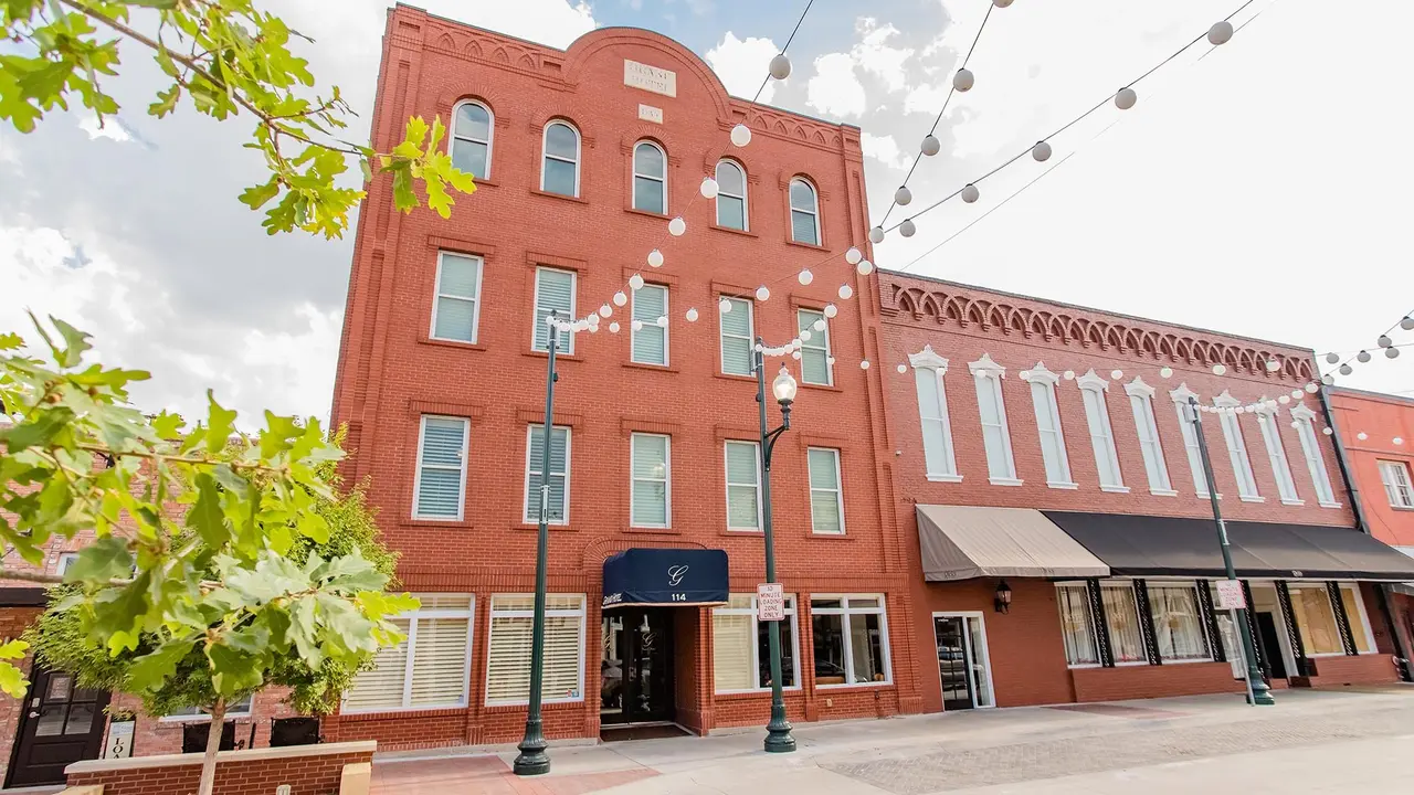 Historic red brick buildings with storefronts on a downtown plaza, strung lights overhead, green foliage visible