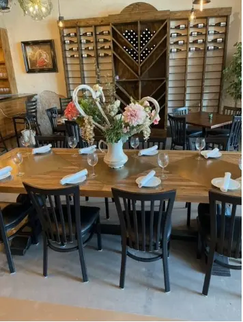 Wooden dining table set with place settings and fresh flowers in a vase, with black chairs and wine storage visible in background