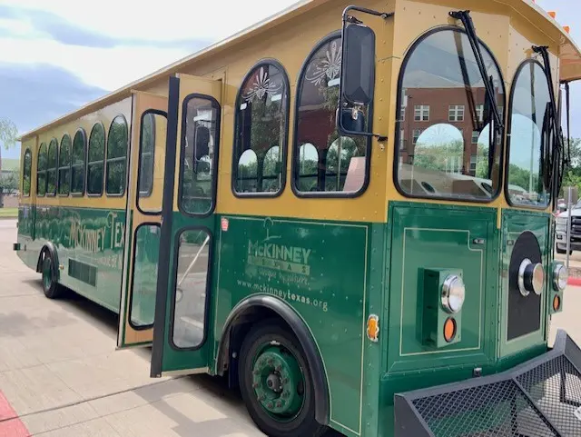 Yellow and green open-air trolley with large windows parked on a paved area with brick building visible in background