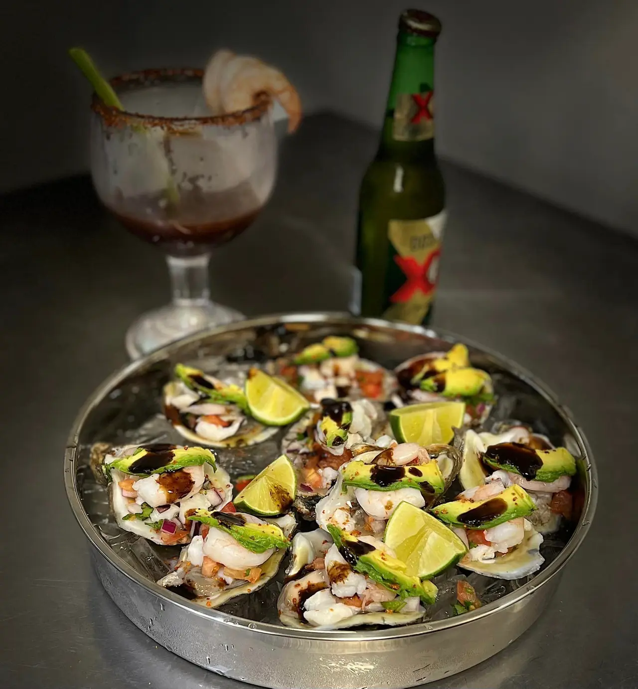 Grilled oysters topped with lime wedges and garnishes arranged on a metal platter, with a cocktail and beer bottle in the background