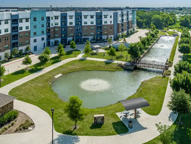 Multi-story residential building with colorful facade overlooking landscaped park featuring pond, green lawns, walking paths, and shade trees