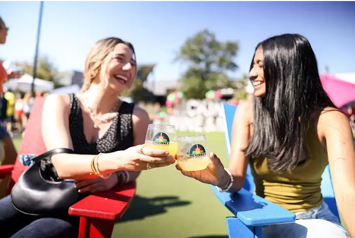 Two women smiling while holding wine glasses at an outdoor festival, with a green lawn and colorful tents in the background