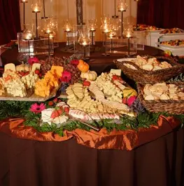 Catered food display with appetizers, cheese, and colorful flowers arranged on tables with warm lighting